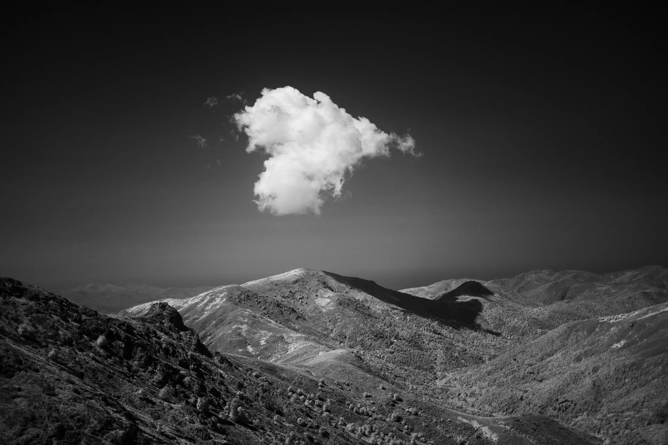 Black and white mountain ridge beneath a single bright cloud in an otherwise dark sky.
