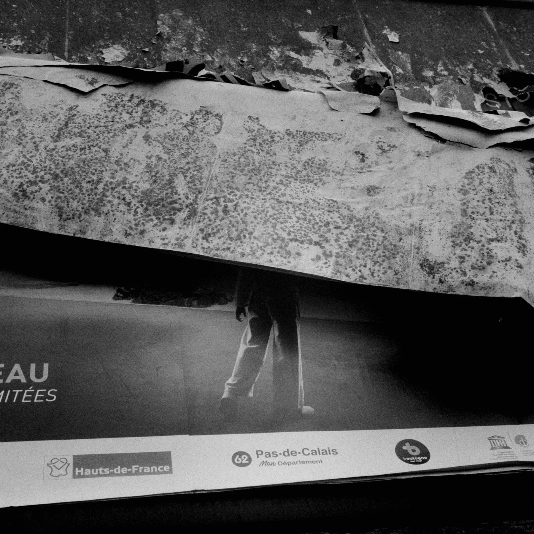 Torn black and white poster revealing the legs of a figure above regional logos.