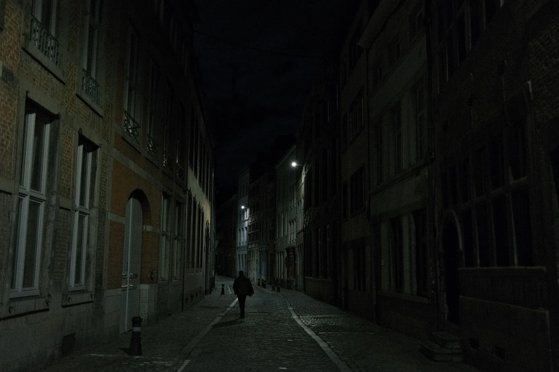 Lone pedestrian walking through a narrow, dimly lit street between old buildings at night.