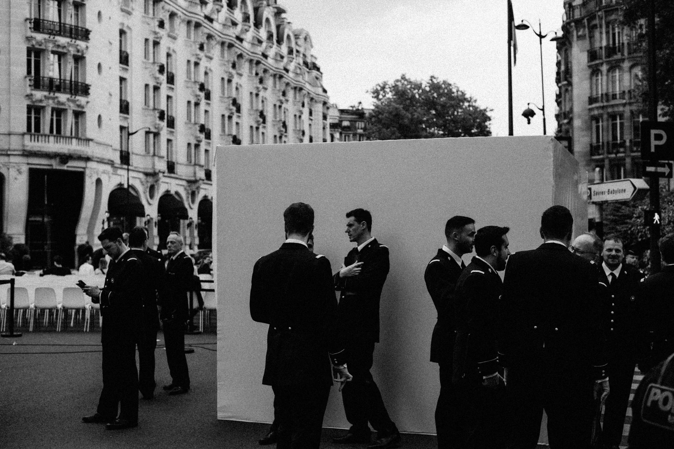 Group of men in dark suits gathered around a white wall on a Paris square.