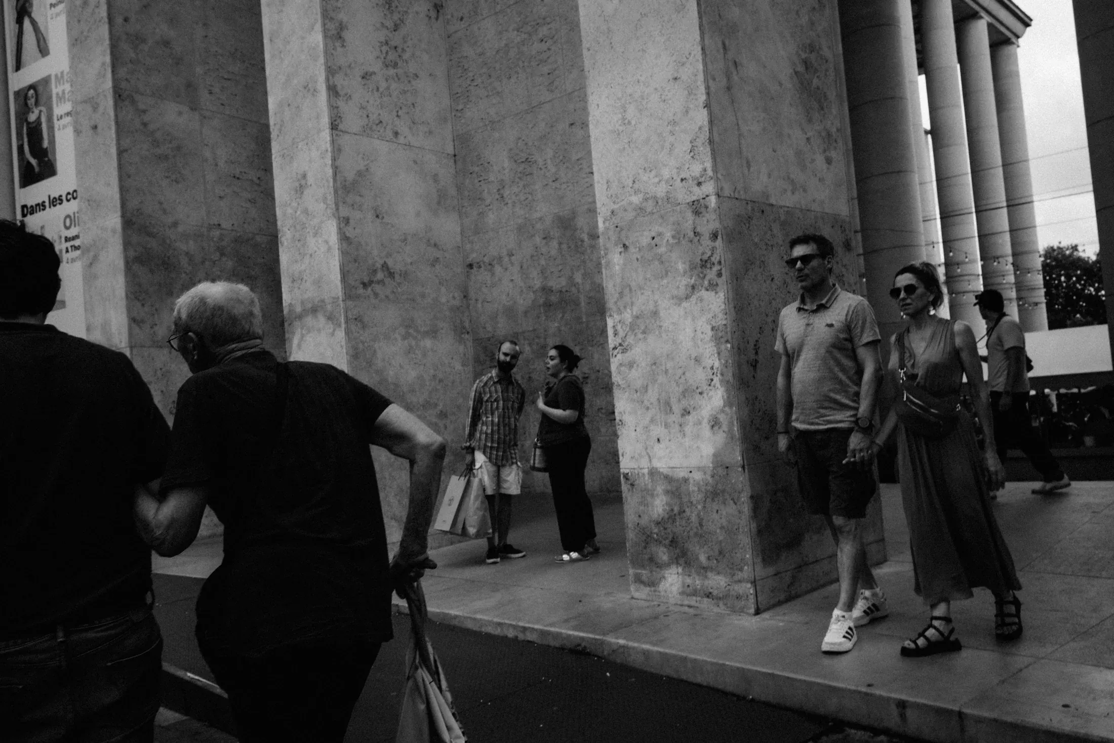 Older man walking past visitors at the base of monumental stone columns in black and white.