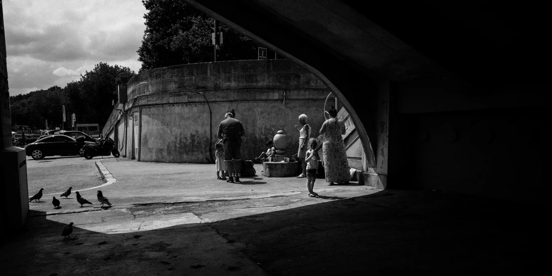 People gathered beneath a concrete arch in a black and white city scene.