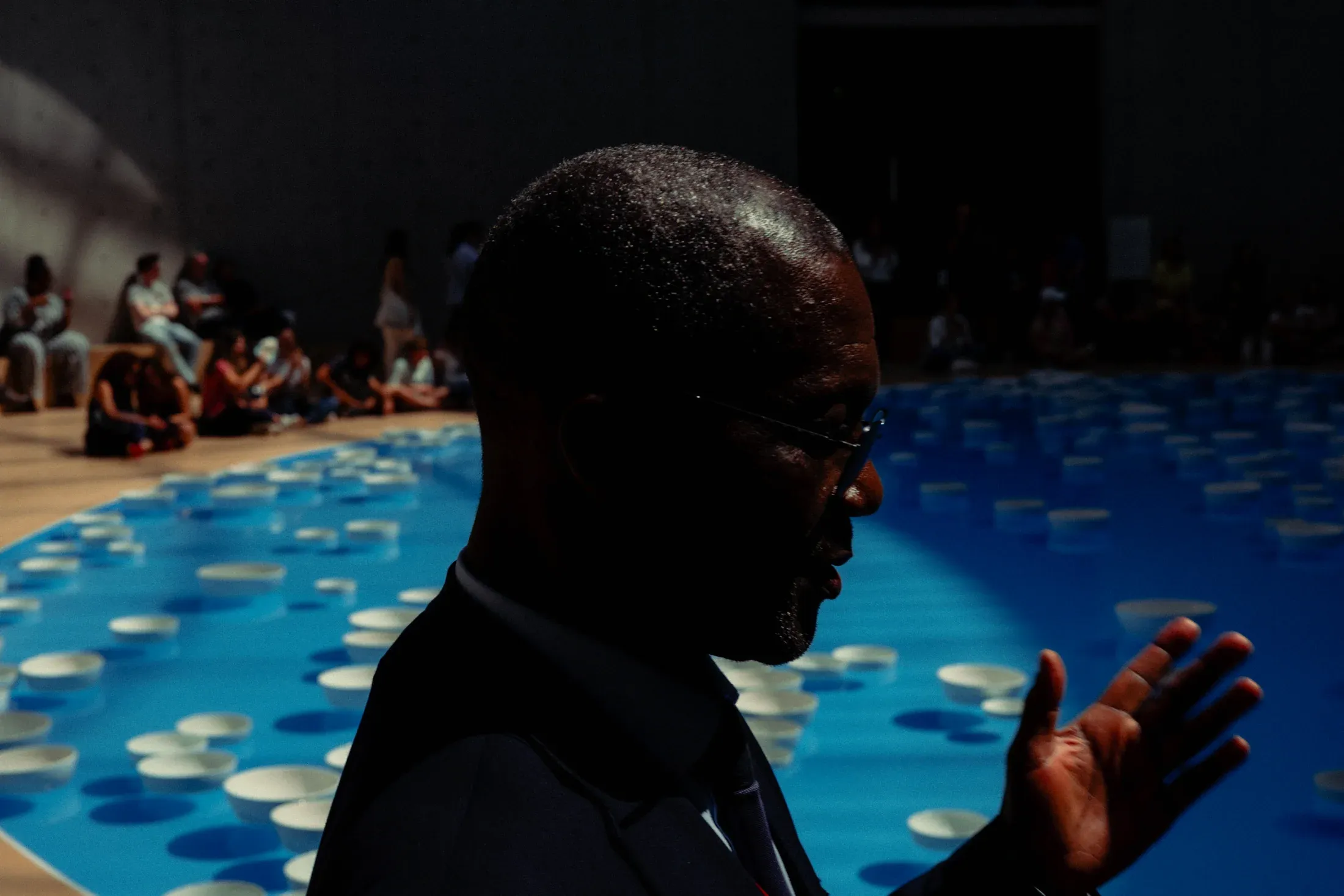 Man in profile gesturing beside a bright blue pool while a seated crowd watches.