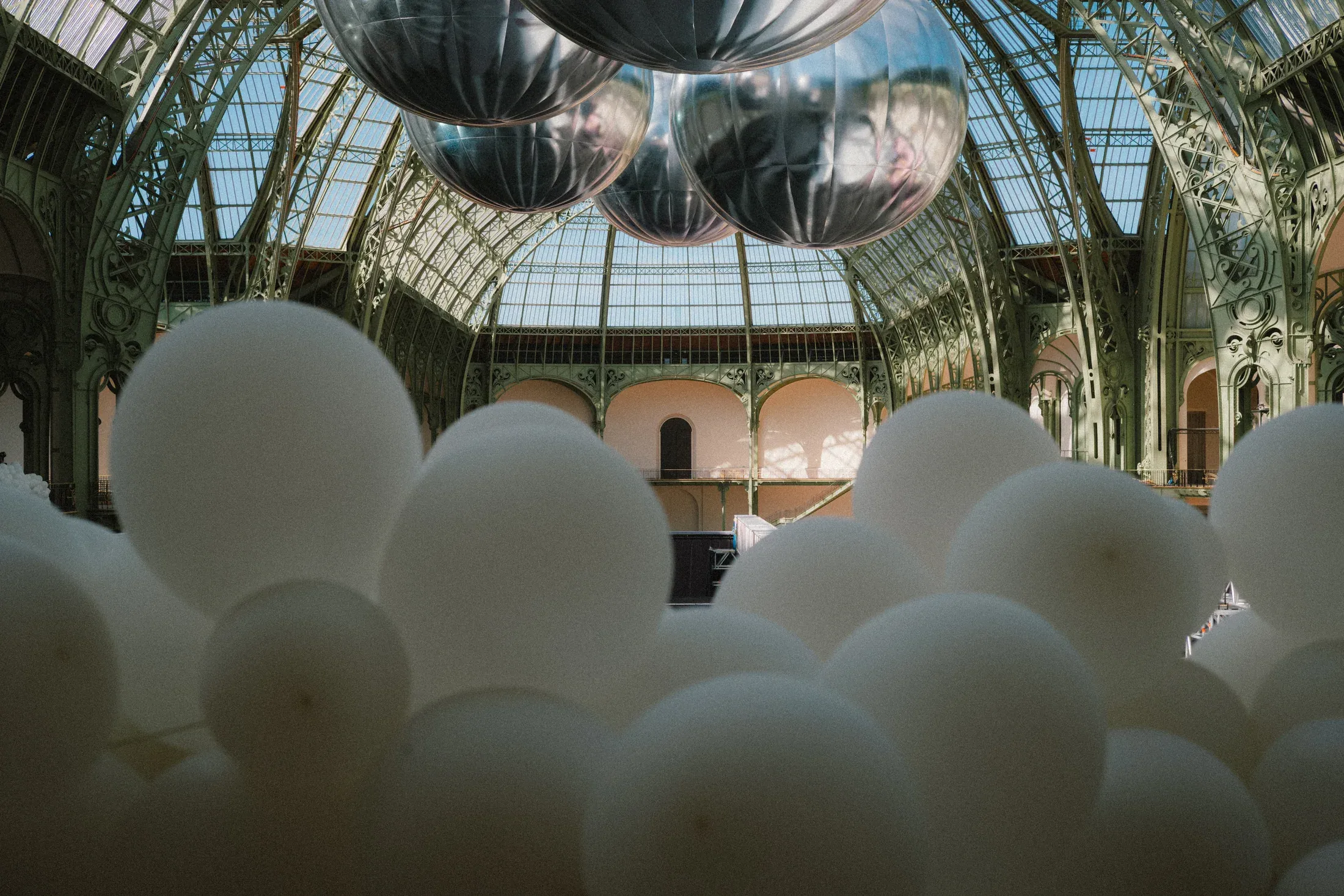 White rounded sculptures and suspended silver forms filling an ornate glass-roofed hall.