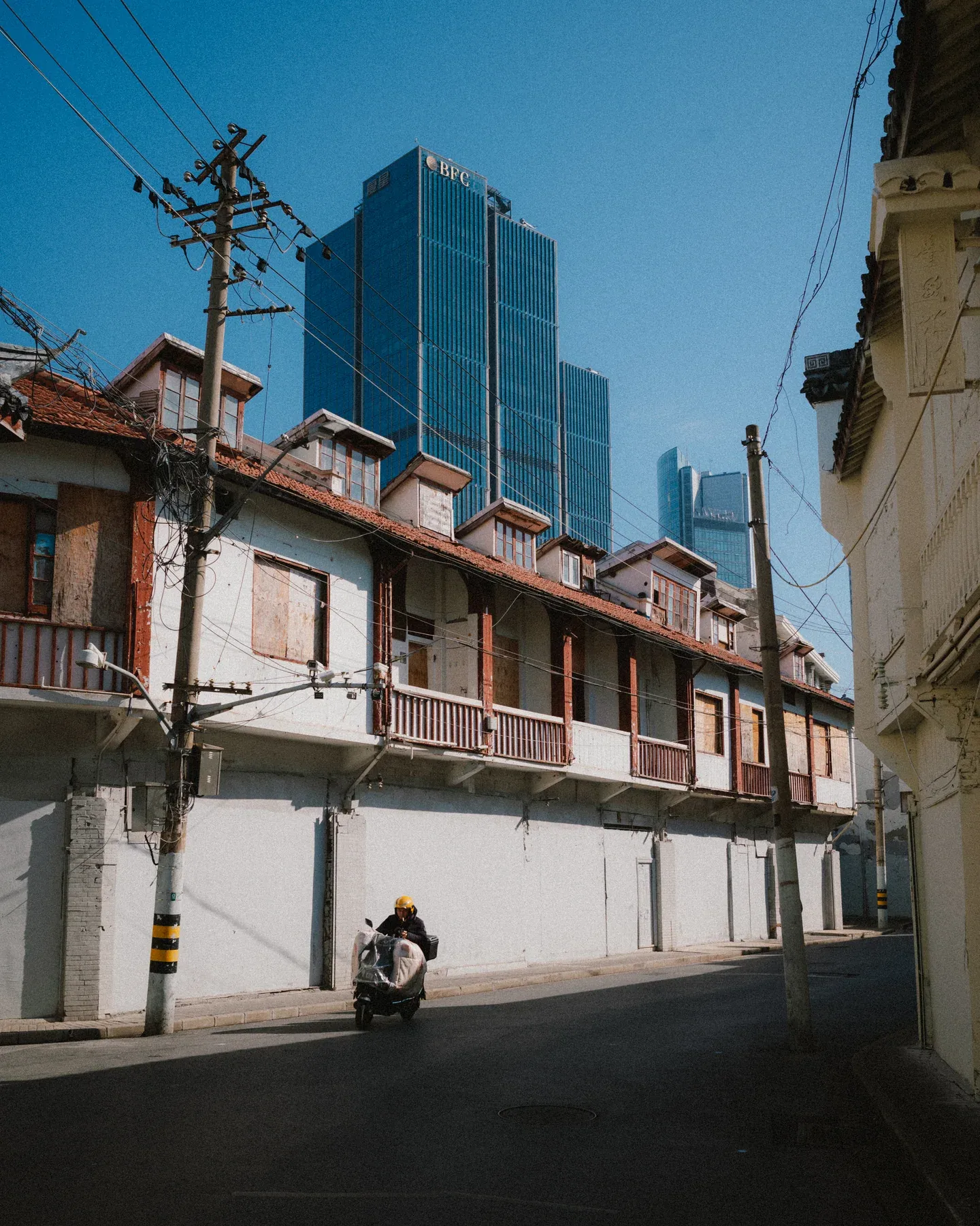 Scooter rider passing old tiled houses beneath blue glass towers and tangled utility wires.