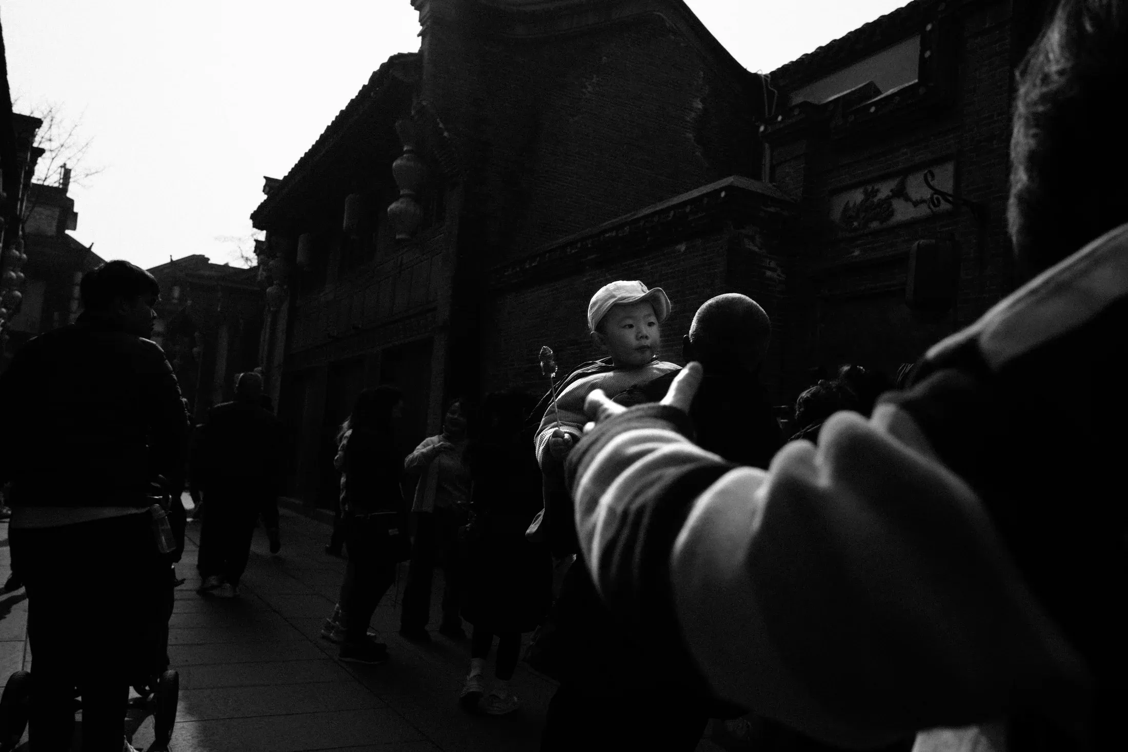 Baby in a sun hat held above a crowd on a shadowed street in black and white.