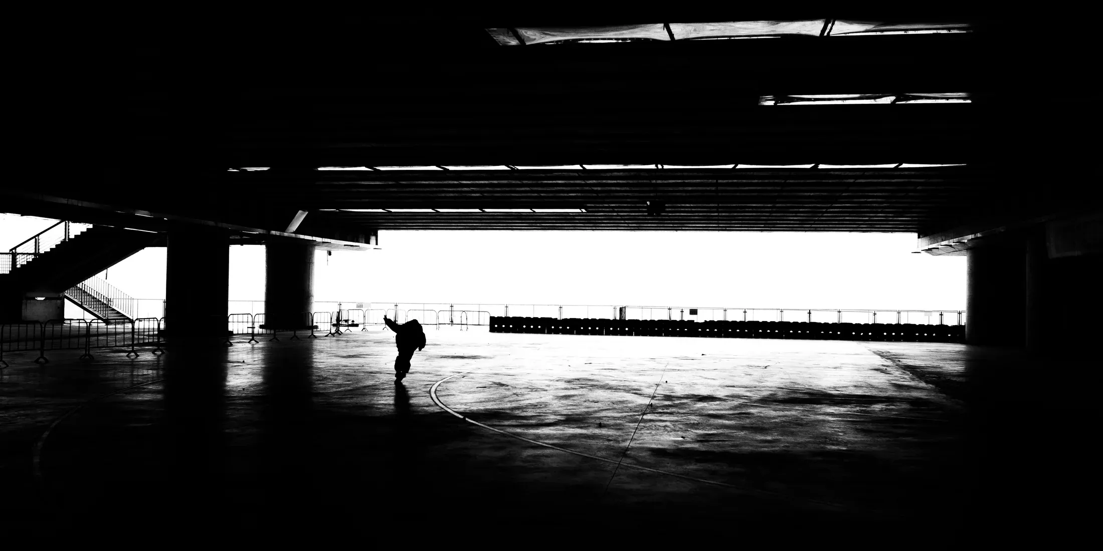 Lone figure crossing a bright strip of ground beneath a vast dark overpass in black and white.