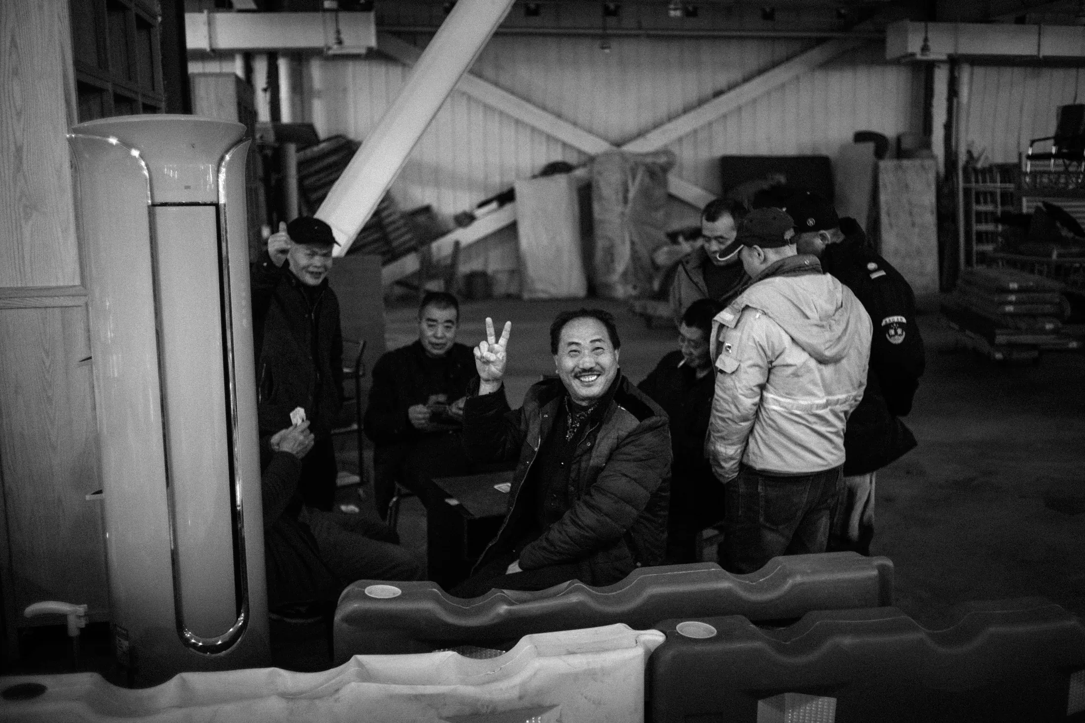 Smiling man giving a peace sign among a group gathered in an industrial workshop.