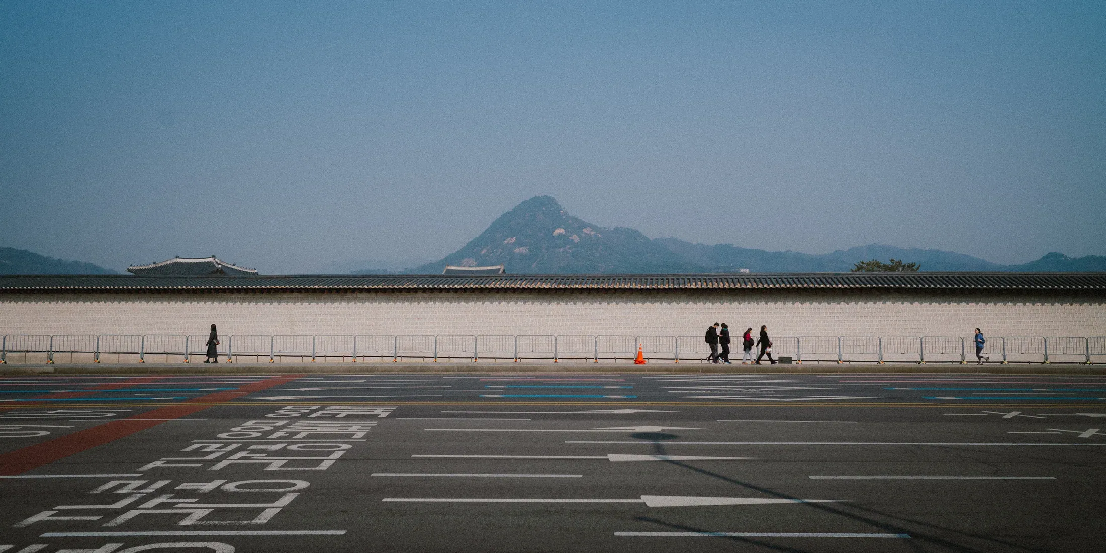 Distant mountain beyond a wide city parking area with small figures crossing the frame.