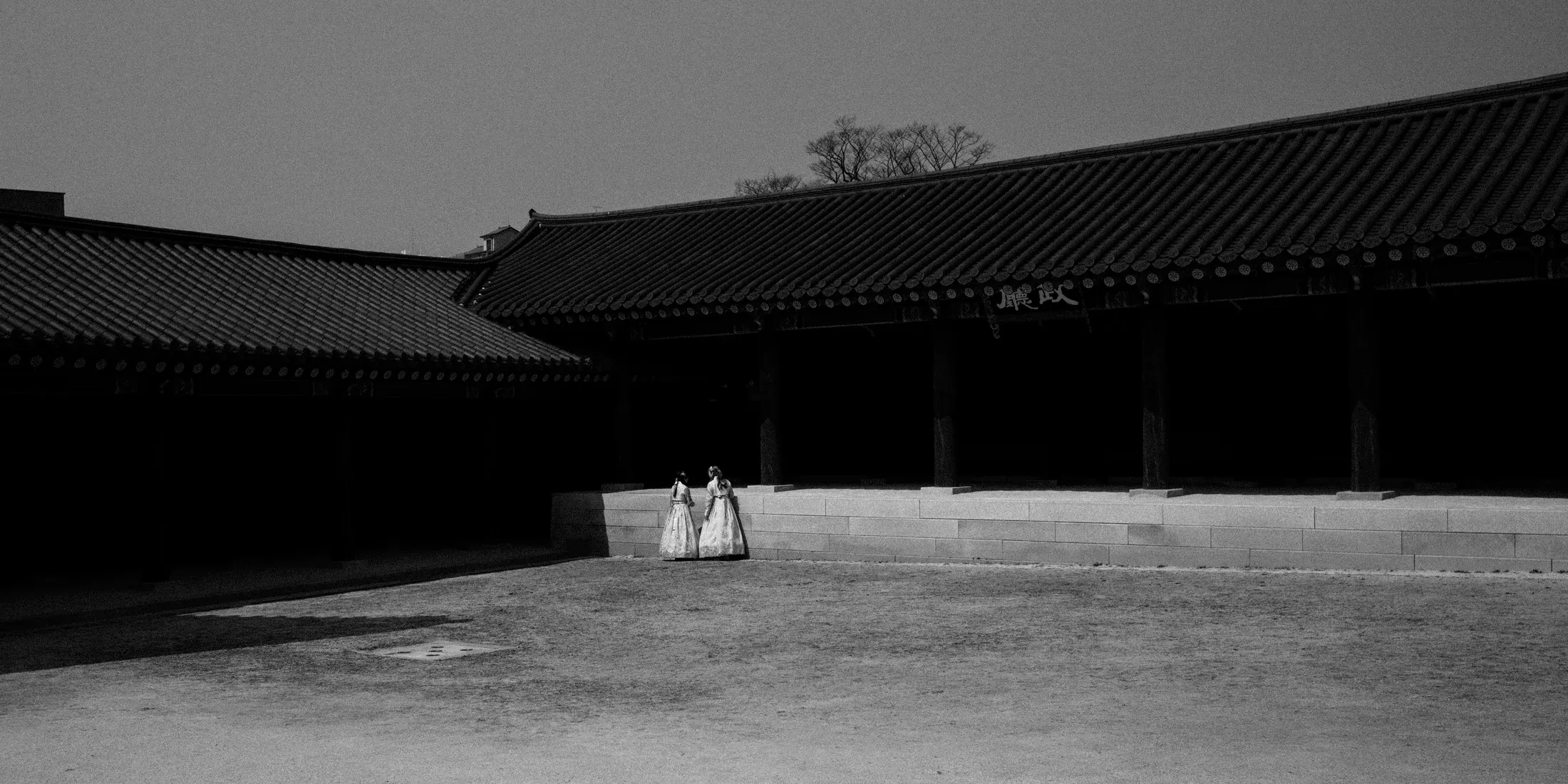 Black and white palace courtyard with two people in traditional dress standing in a bright open yard.