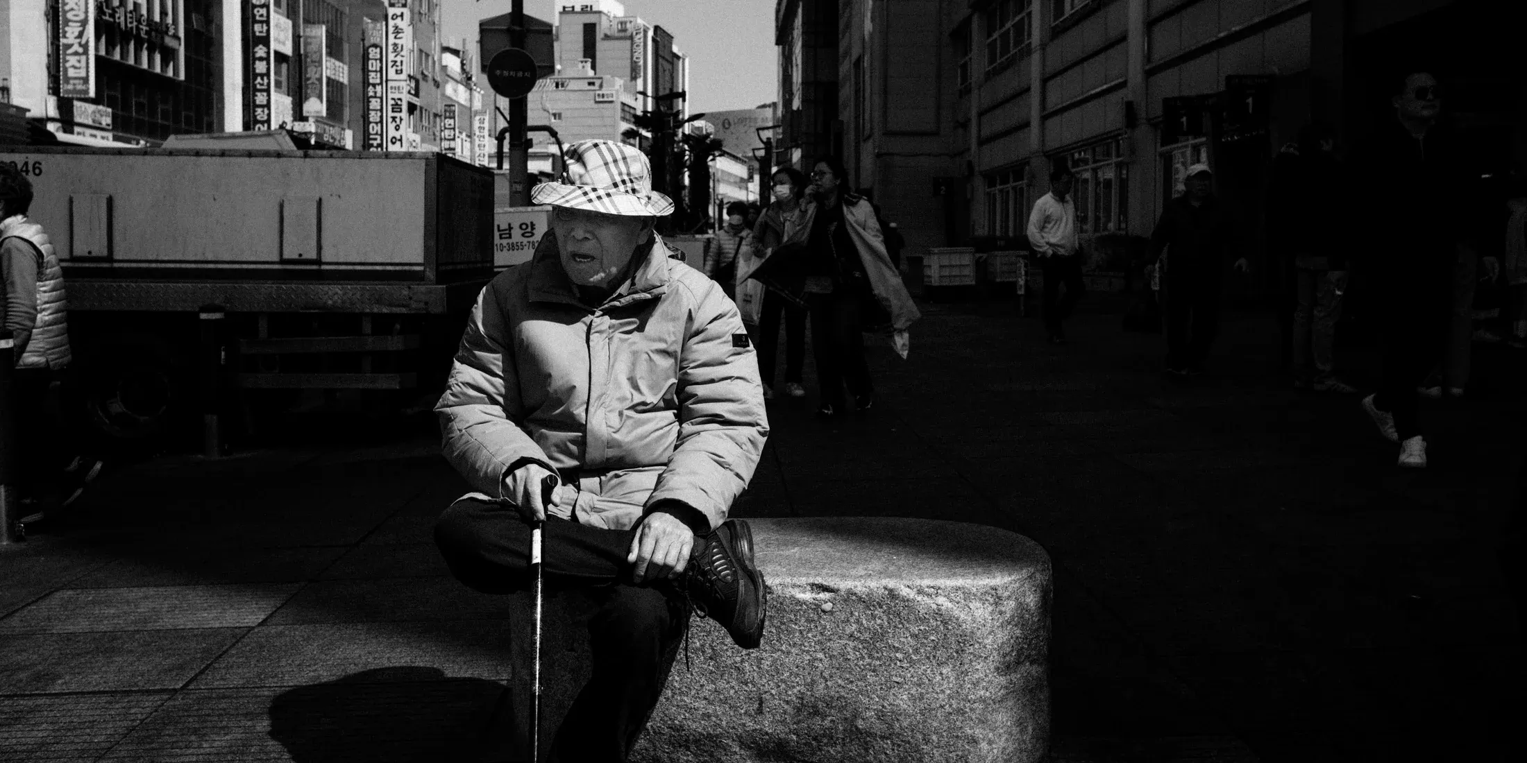 Black and white street portrait of an older person seated in shade with traffic and pedestrians behind.
