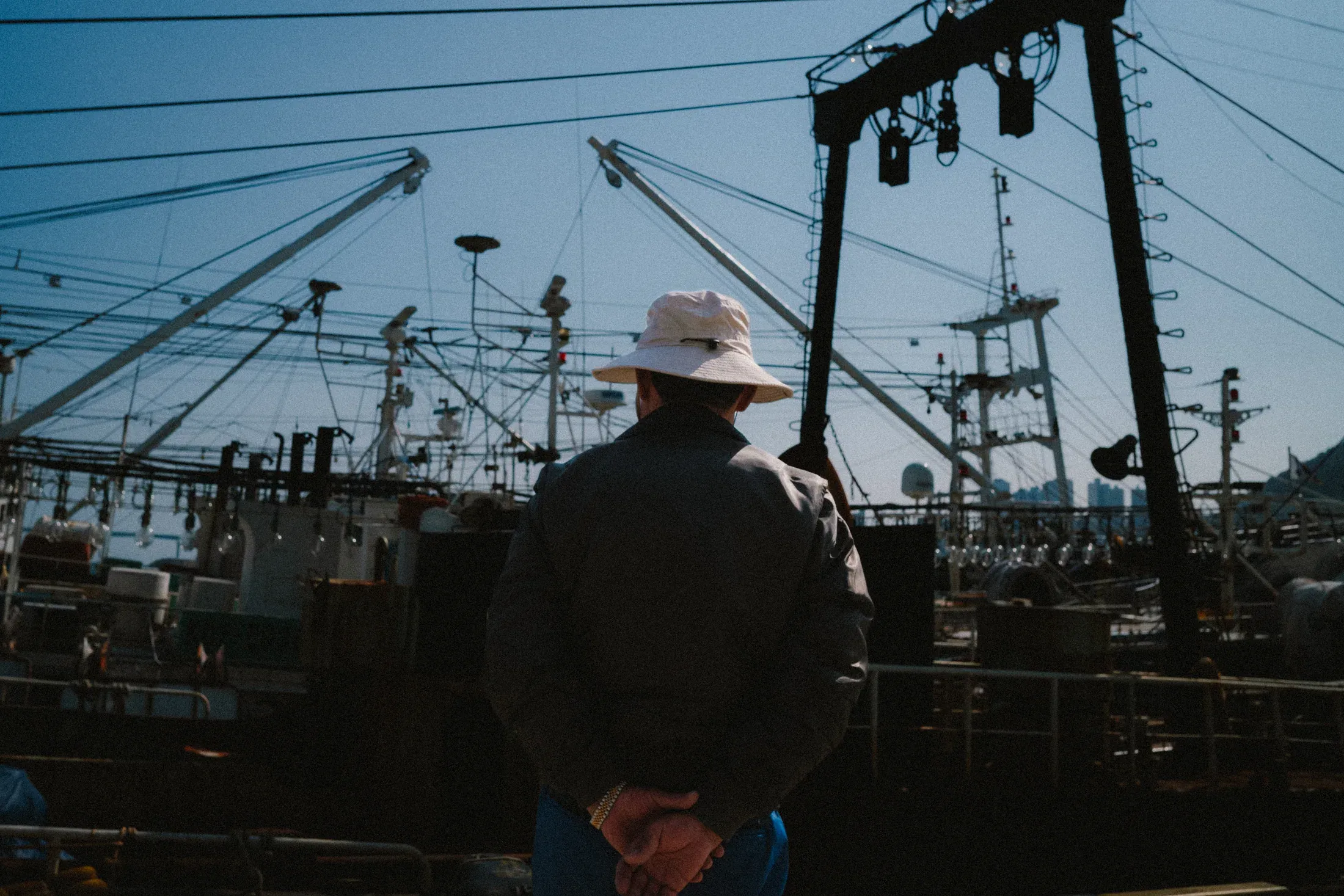 Person in a brimmed hat standing with hands behind their back among wires, poles, and industrial fixtures.