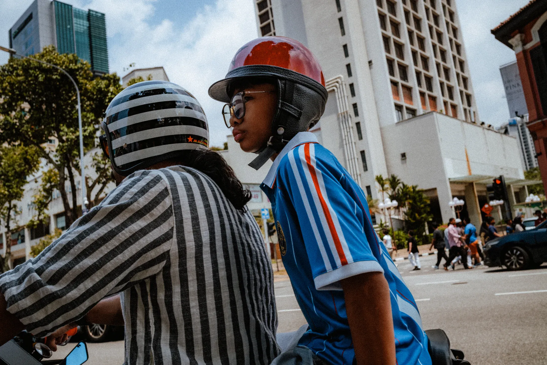 Two scooter riders in striped shirts and helmets stopped on a sunny Singapore street.