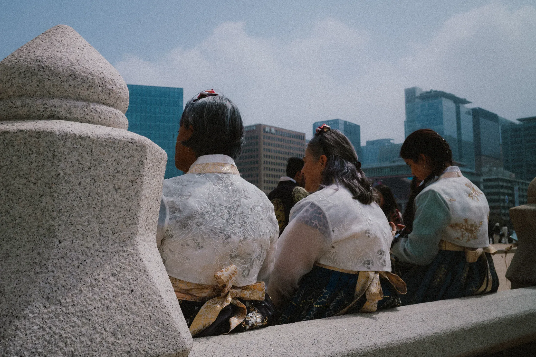 People in pale traditional clothing sitting on a stone edge with glass buildings behind them.