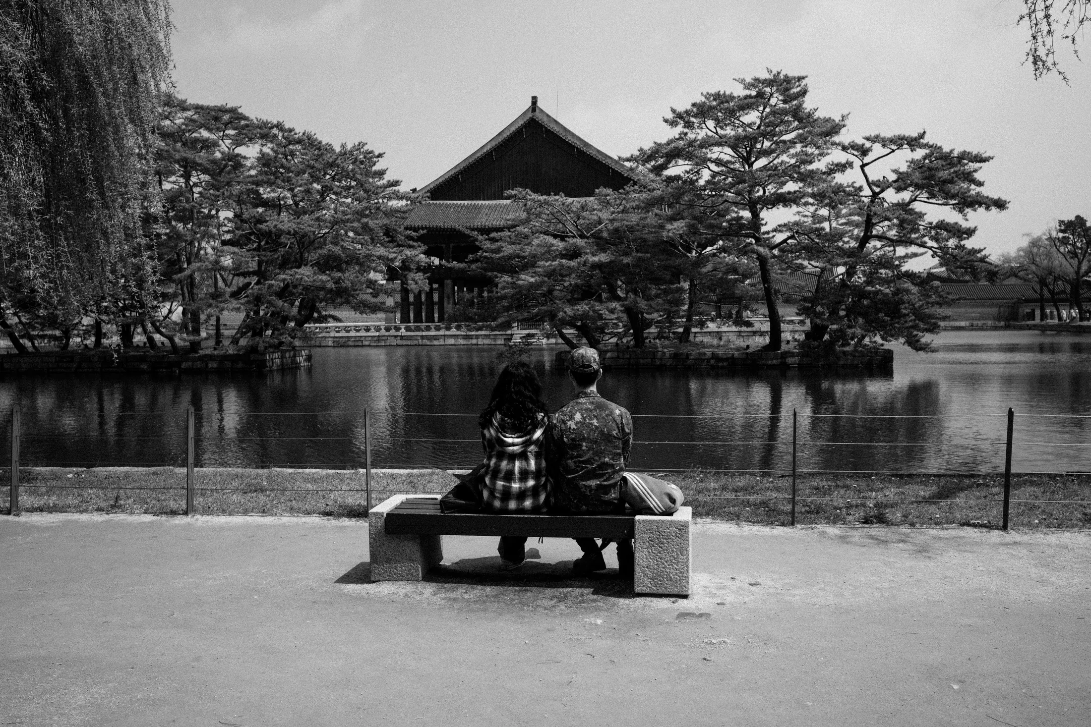 Black and white pond scene with two people seated on a bench facing a pavilion and trees.