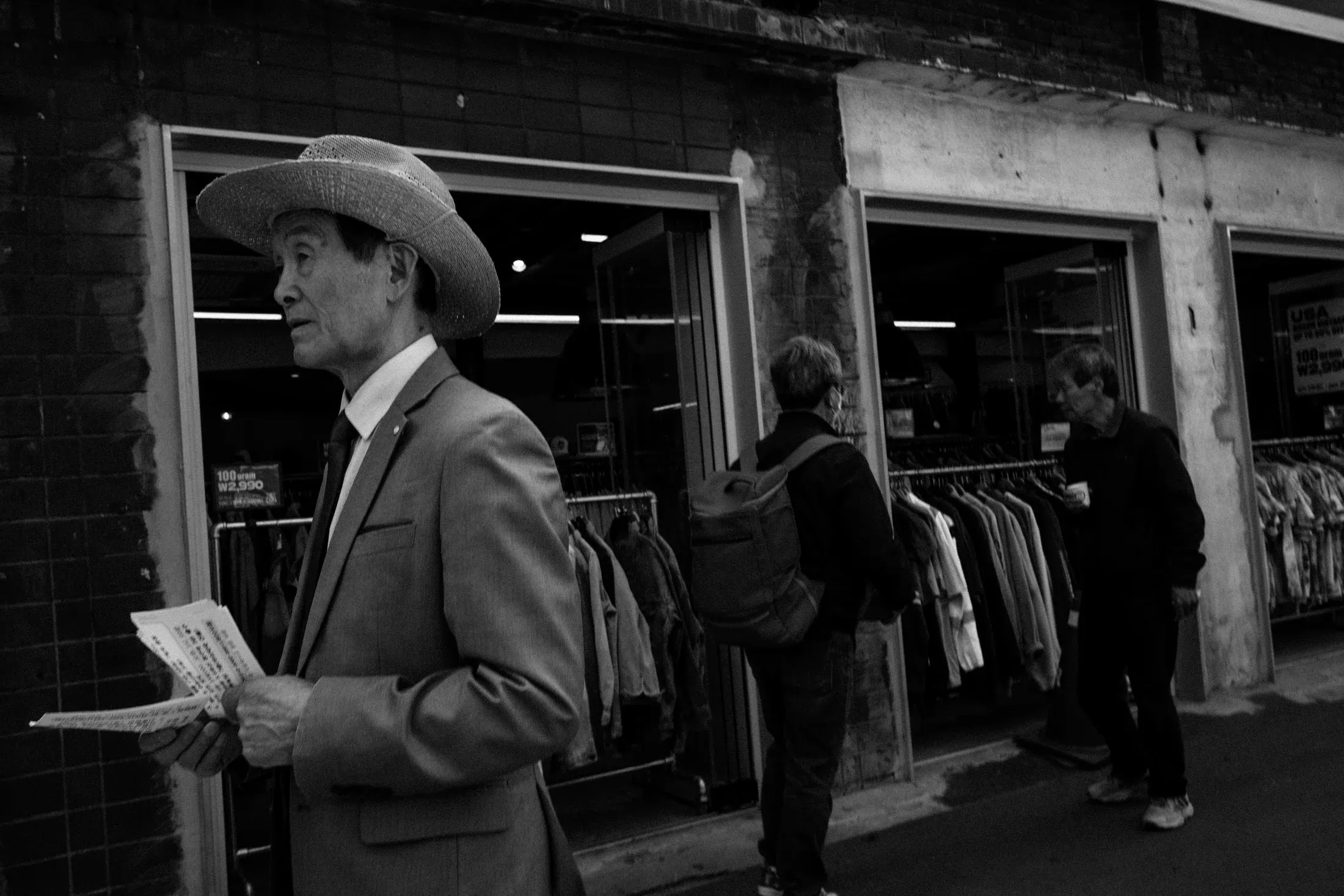 Black and white street scene with a man in a cowboy hat holding papers outside shopfronts.