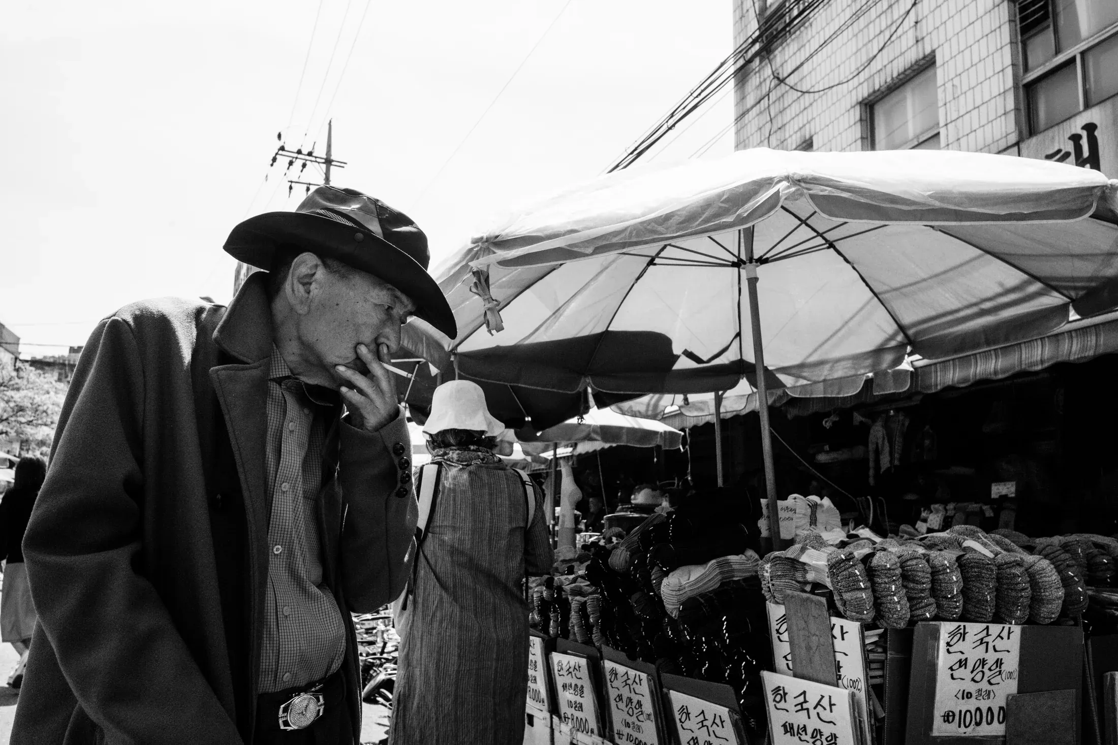 Black and white market scene with an older man smoking beneath umbrellas and hanging wires.