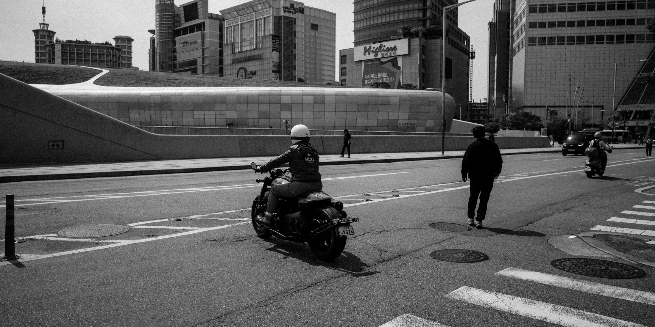 Black and white city road with motorbikes and pedestrians near a large curved metallic building.