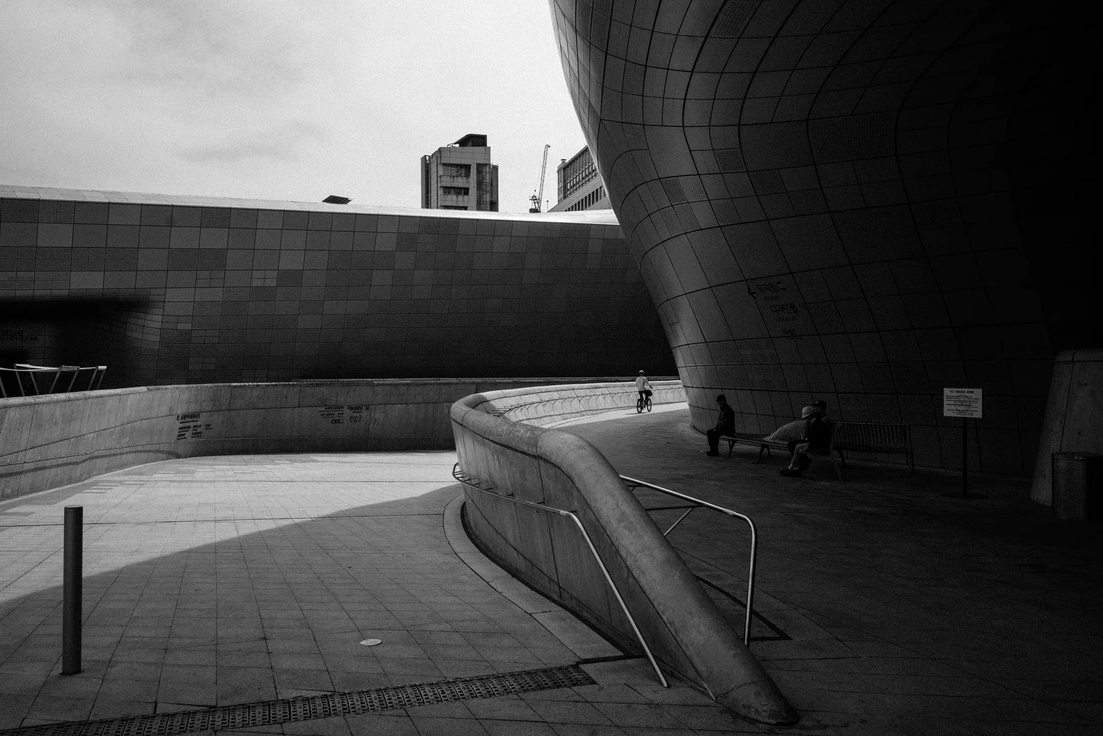 Black and white architectural plaza with curving metal surfaces, stairs, and a small seated figure.