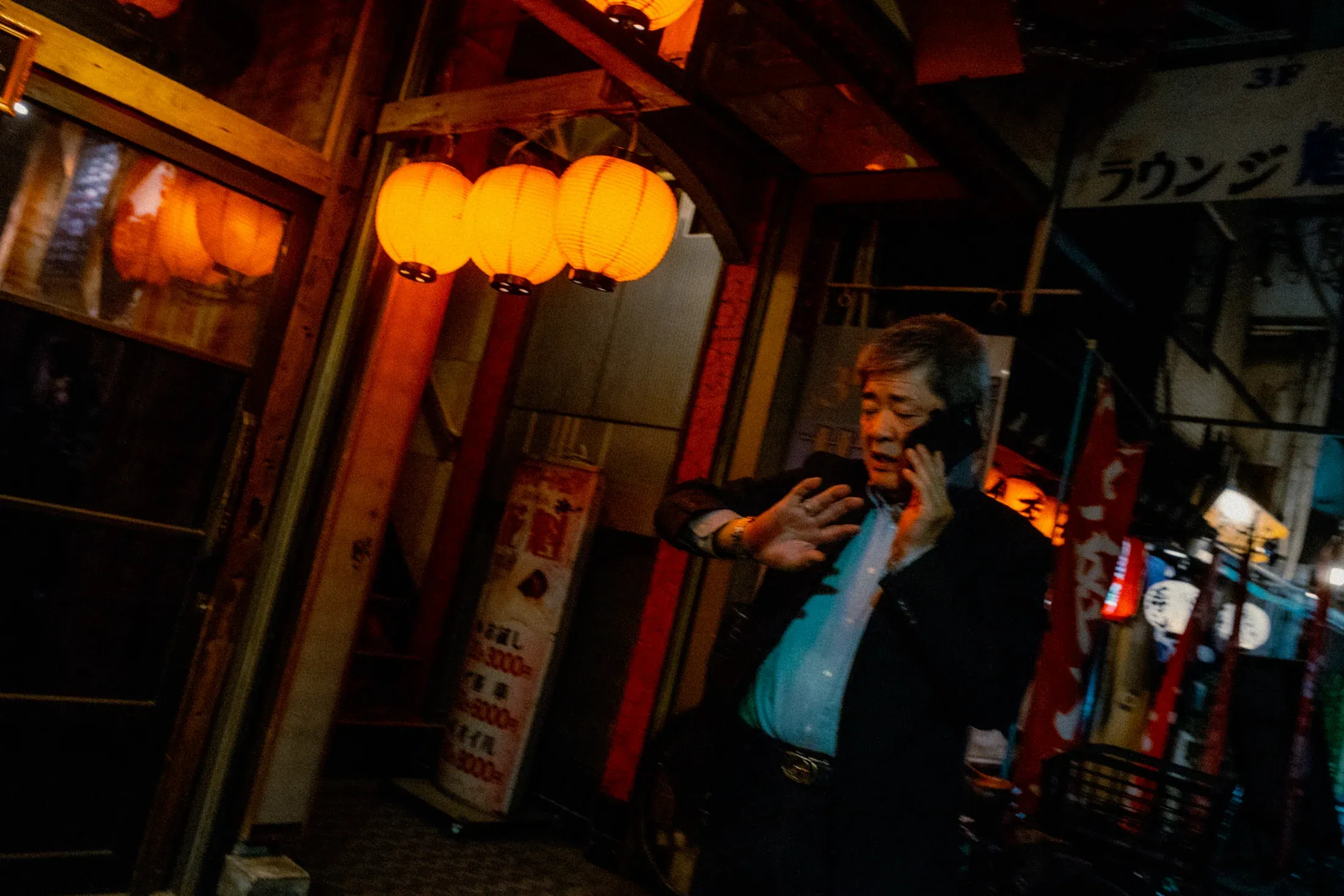 Man standing beneath warm red lanterns outside a narrow night storefront.