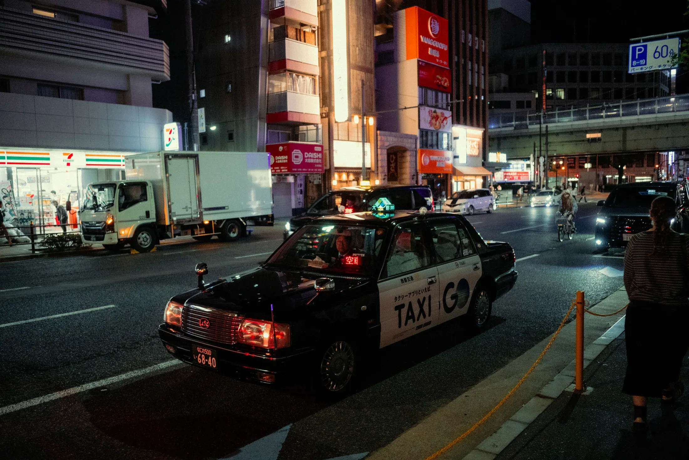 Night street with a taxi stopped near lit storefronts and passing traffic.