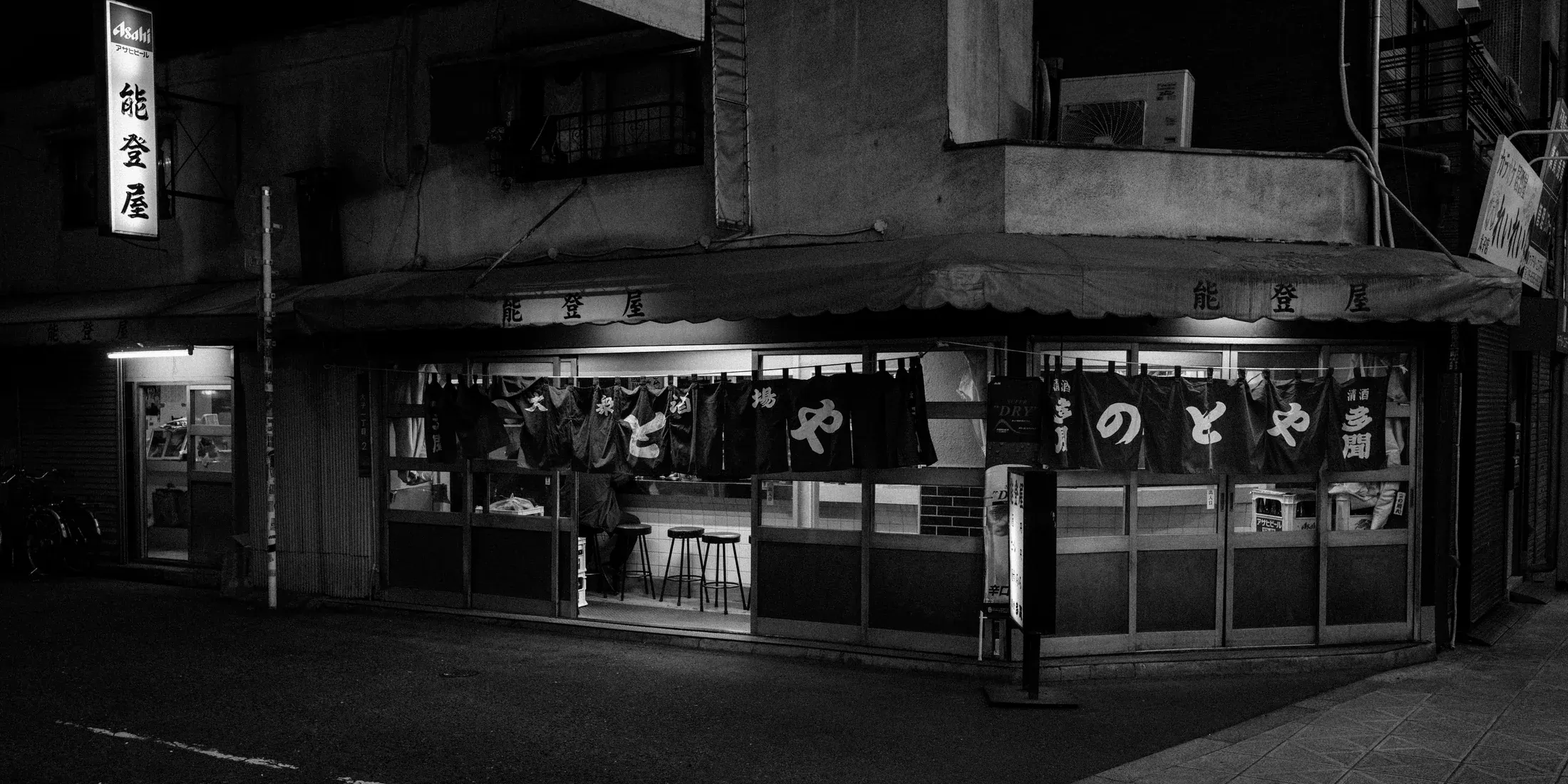 Black and white night storefront with a single figure standing at the corner.