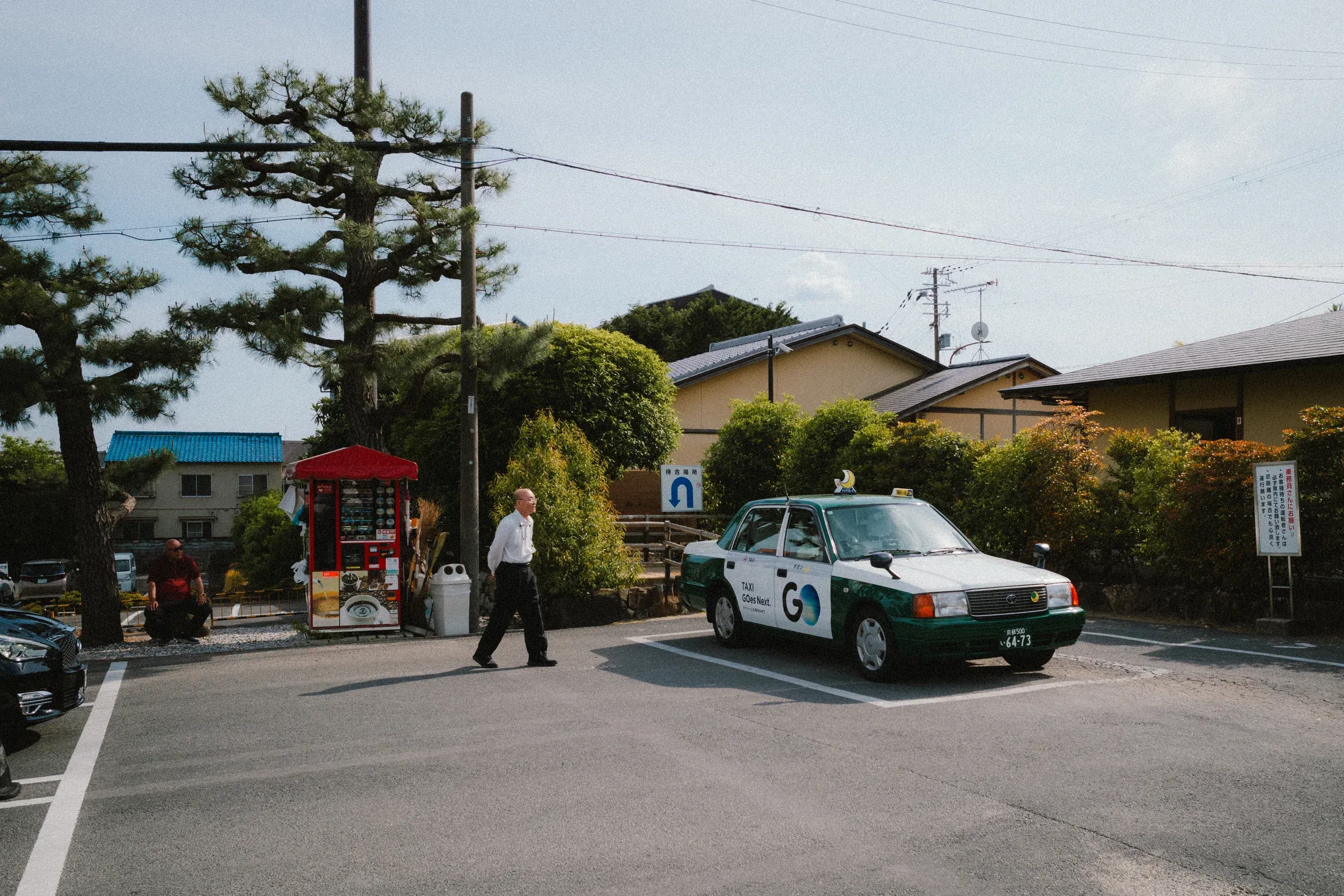White taxi and a lone pedestrian beside a small roadside kiosk in bright afternoon light.