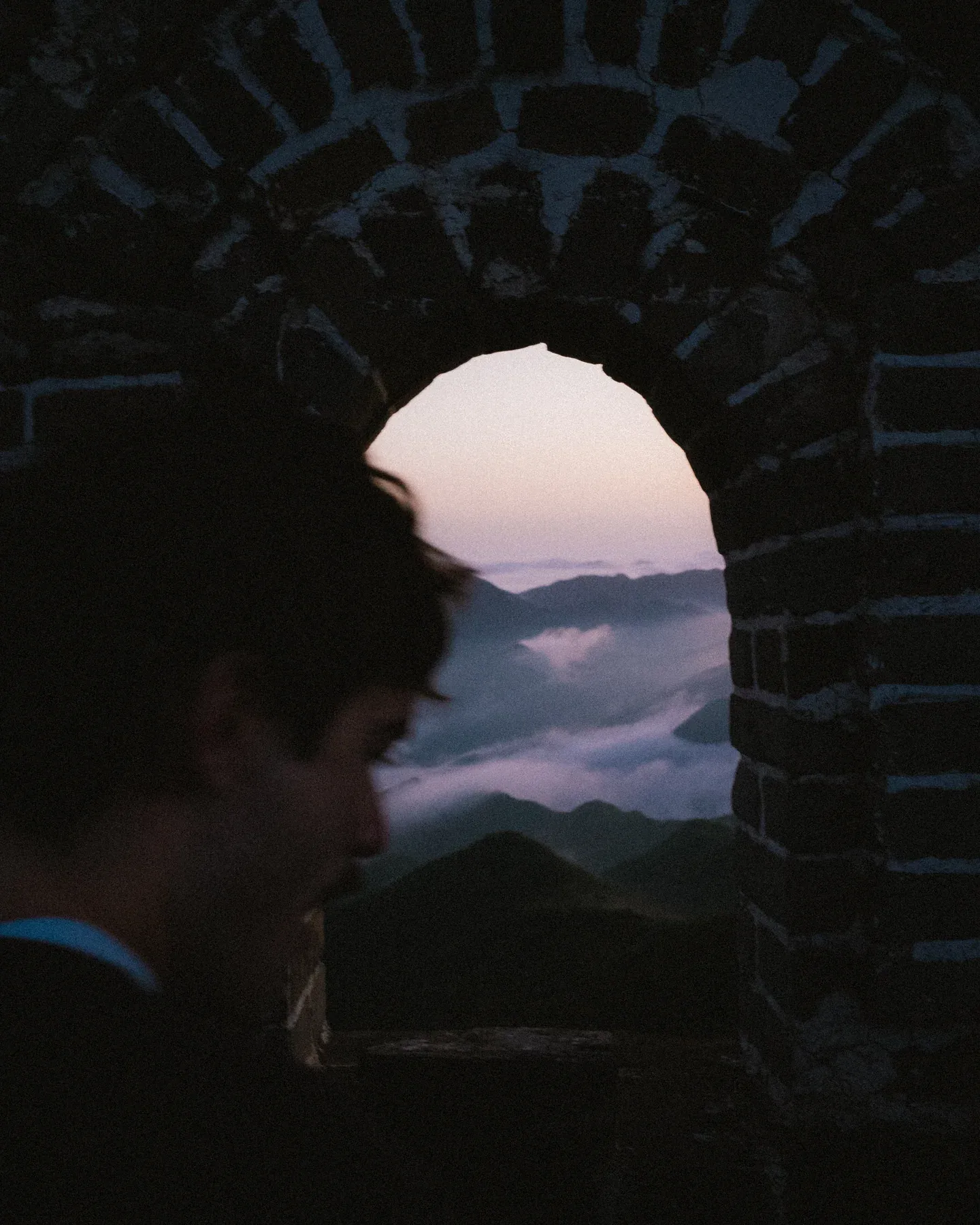 Profile of a person framed in a dark stone archway looking toward distant mountains.