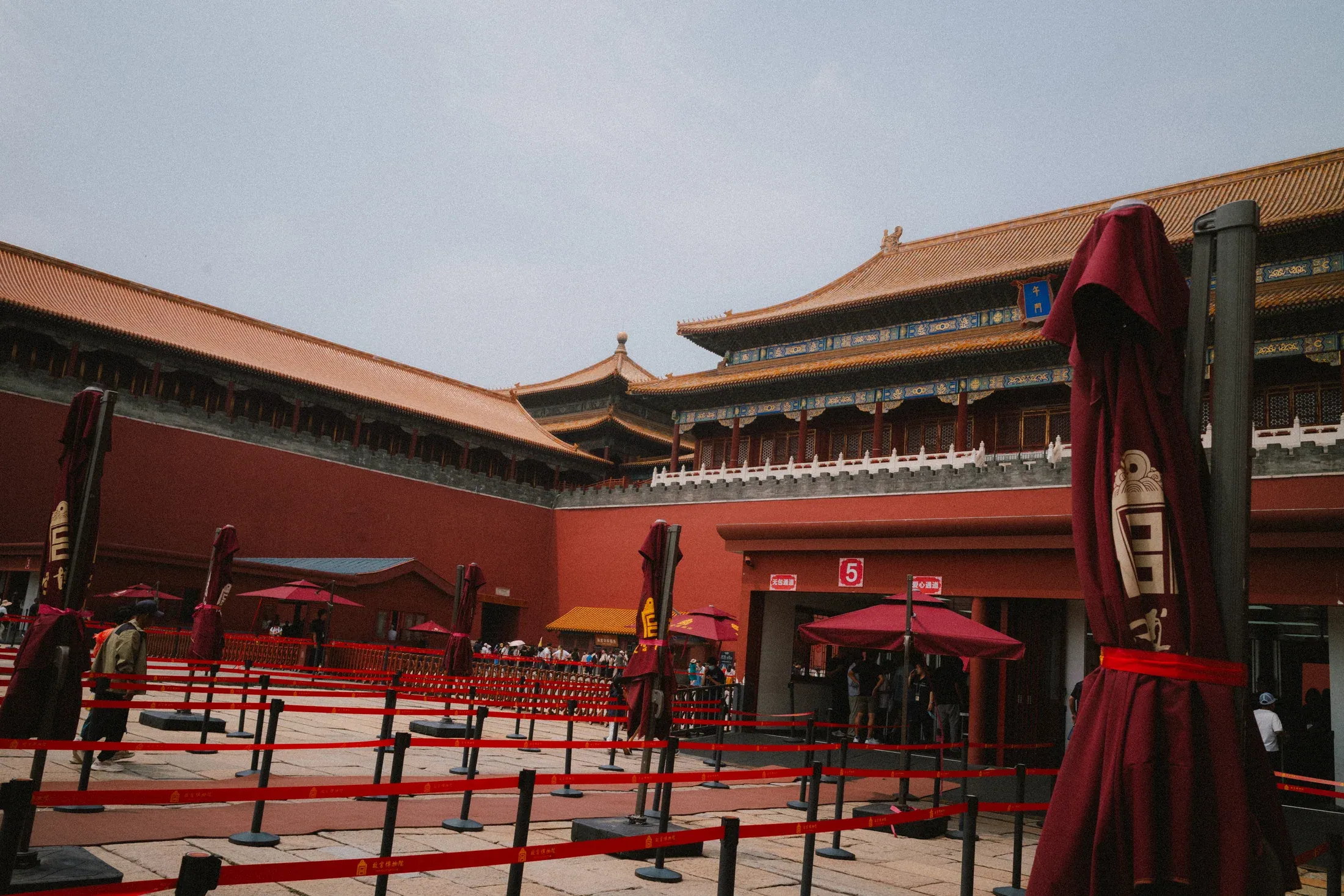 Red palace courtyard with closed umbrellas and people moving through the sun.