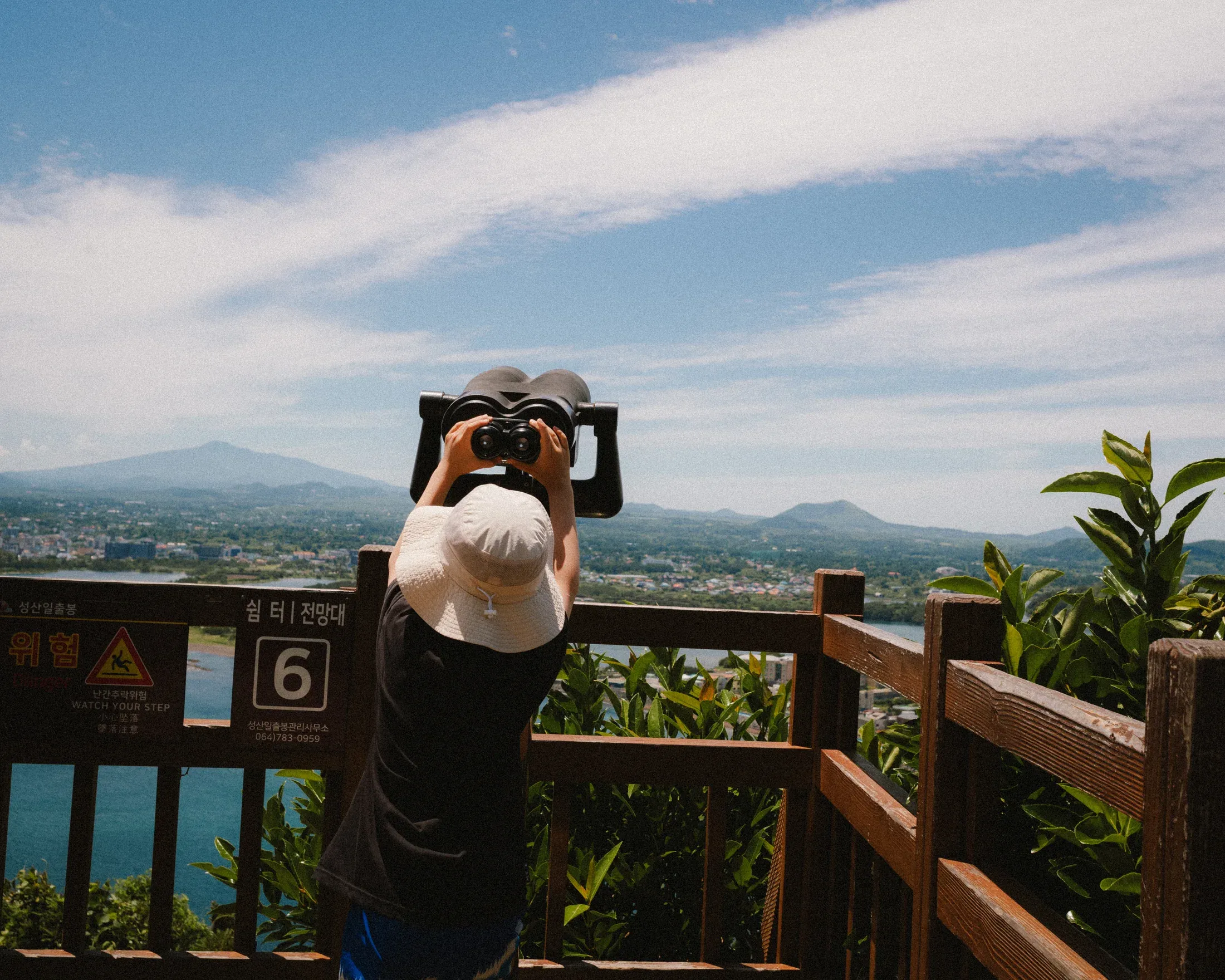 Visitor in a sun hat looking through binoculars toward a coastal town and distant hills.