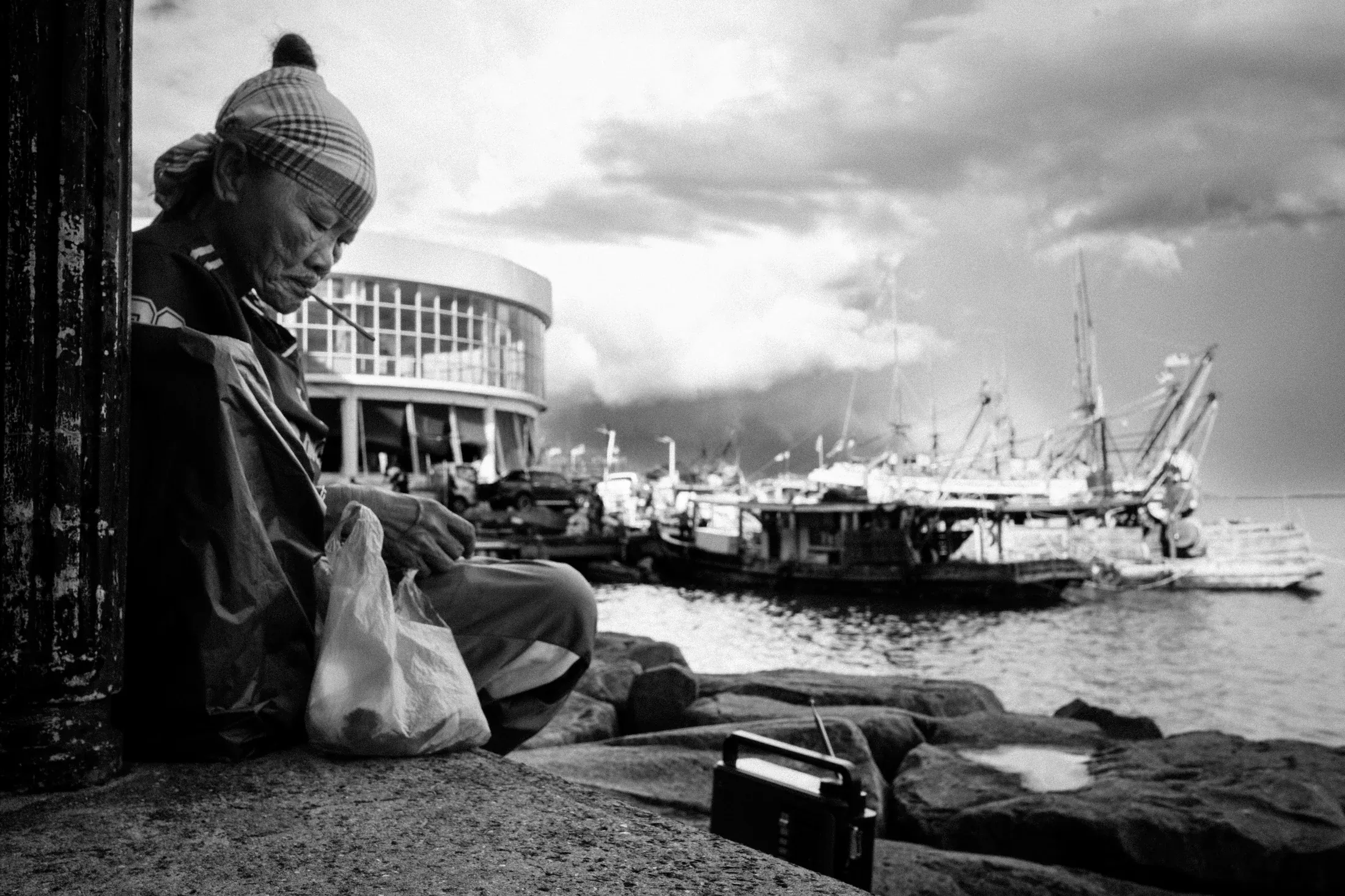 Woman sitting on a harbour wall beside her bags, with boats and cranes beneath a cloudy sky.