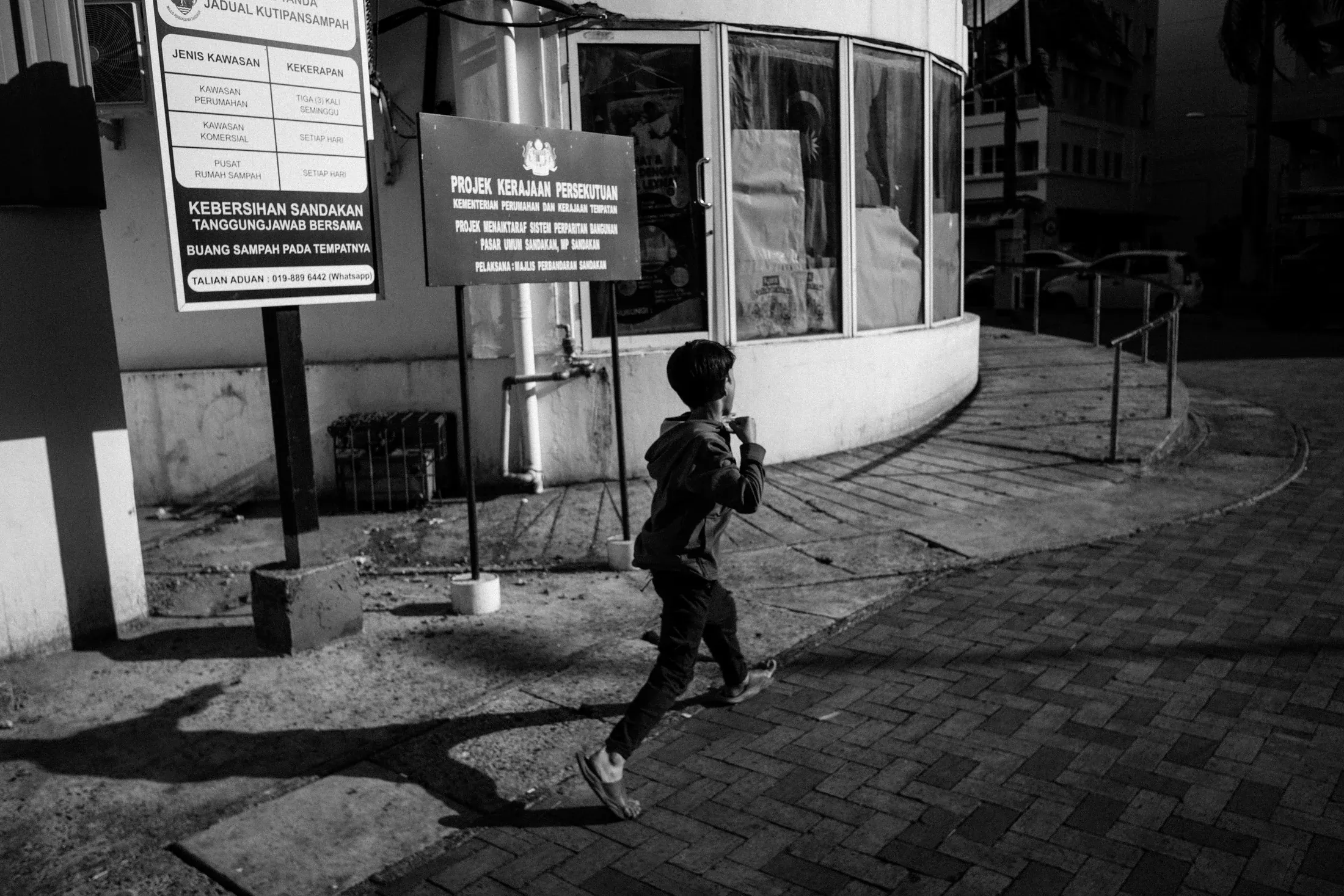 Child running past municipal signs and a curved building in strong black and white light.