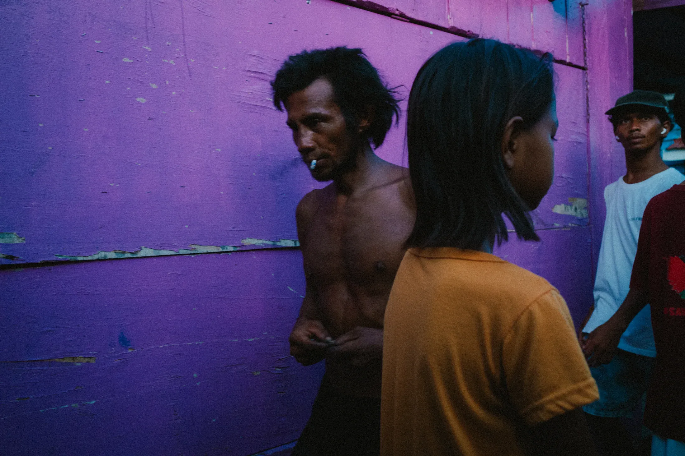 Shirtless man and passing shoppers against a vivid purple market wall.