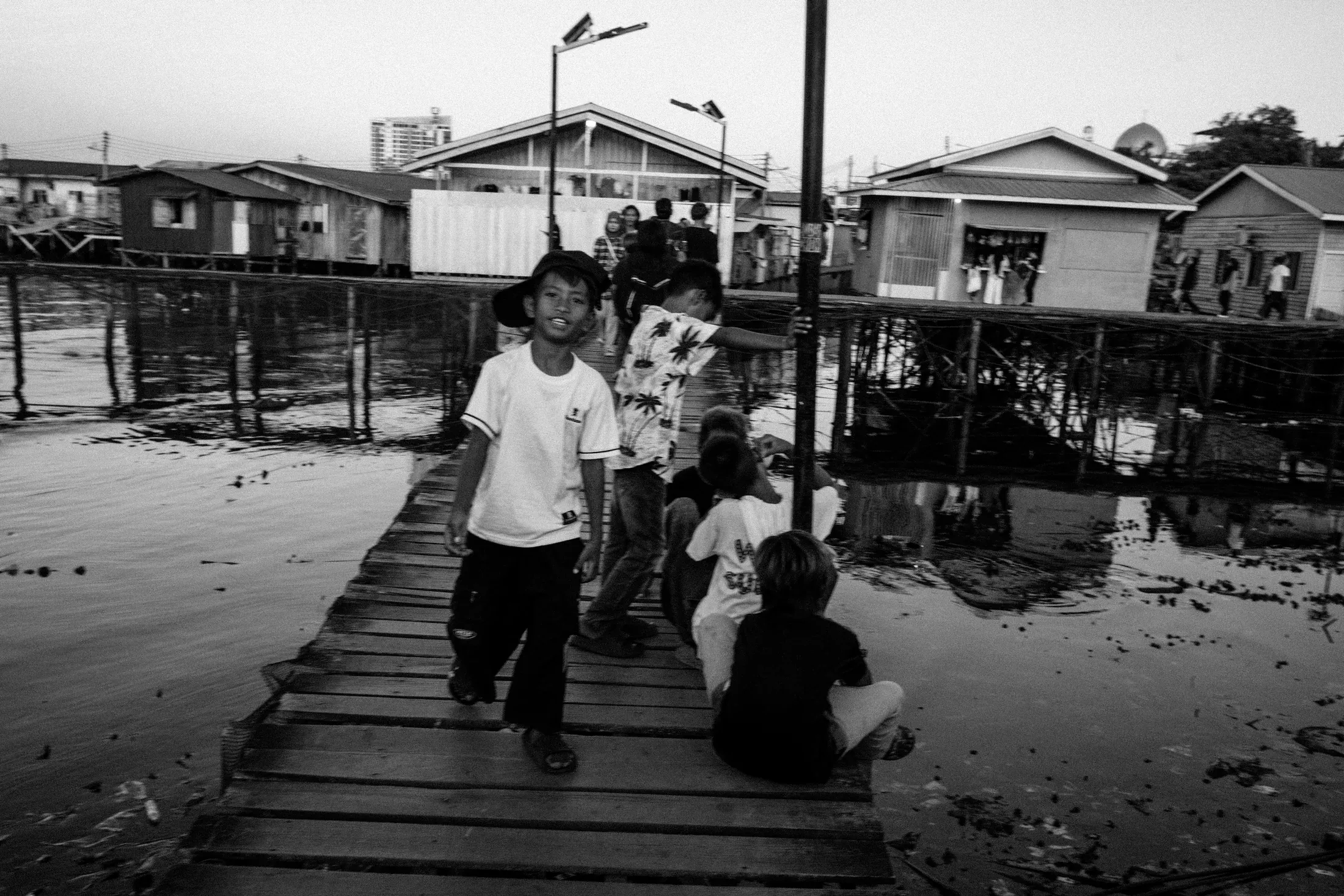 Black and white view of residents walking along a narrow boardwalk between stilt houses.