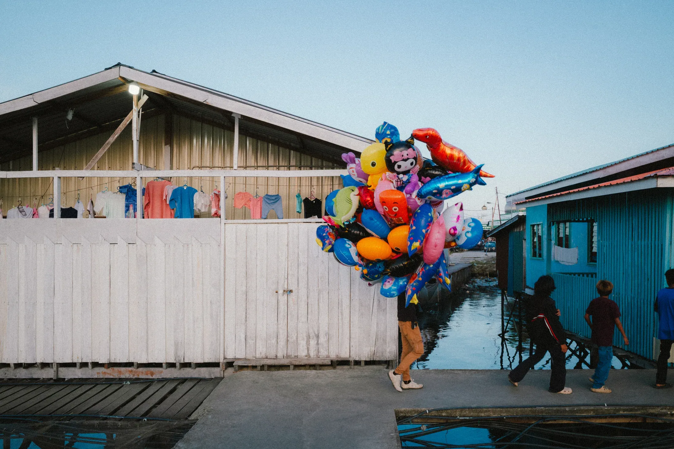 Balloon seller carrying a bright cluster past blue houses and seated children.