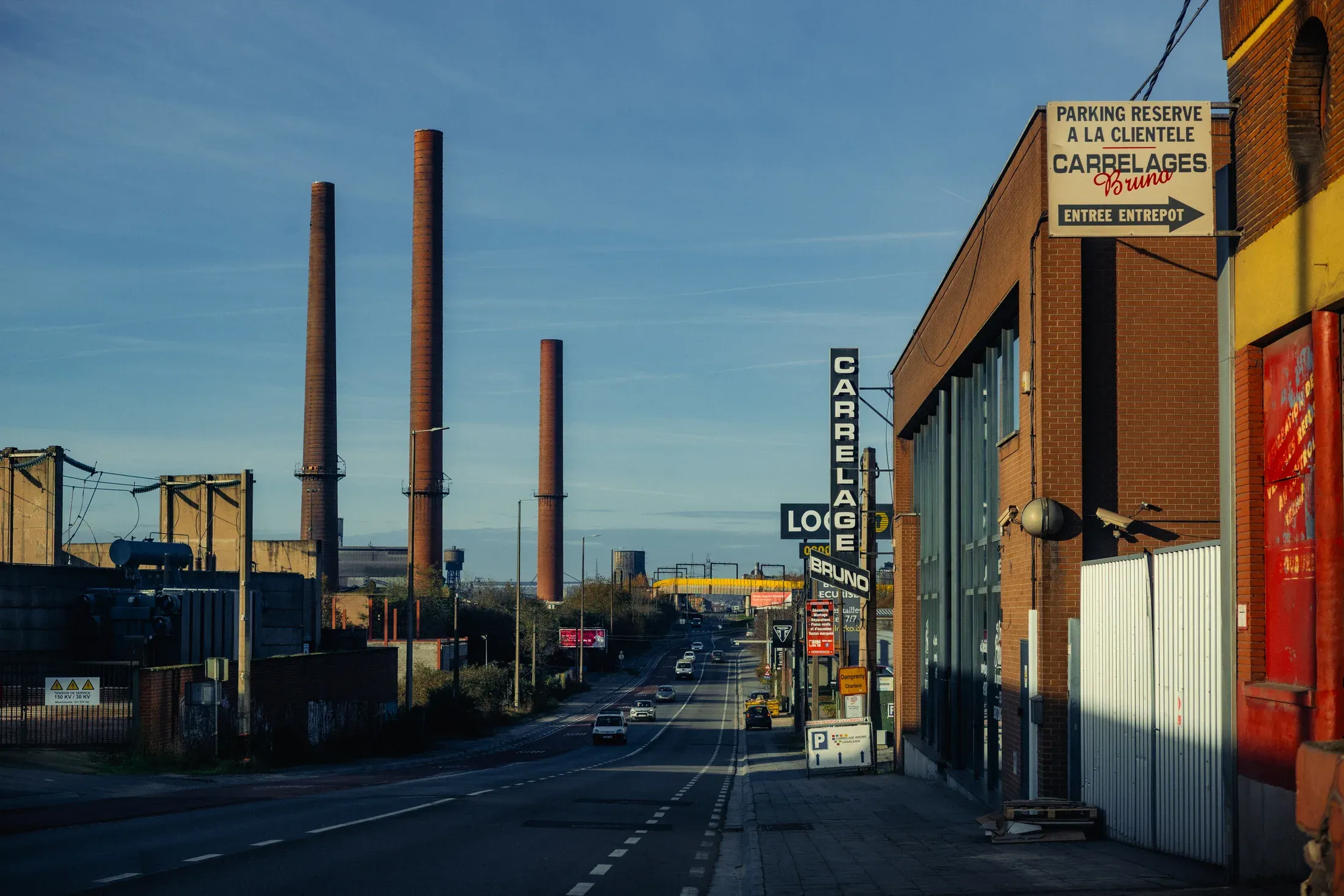 Industrial street lined with brick workshops, signs, and two tall smokestacks.