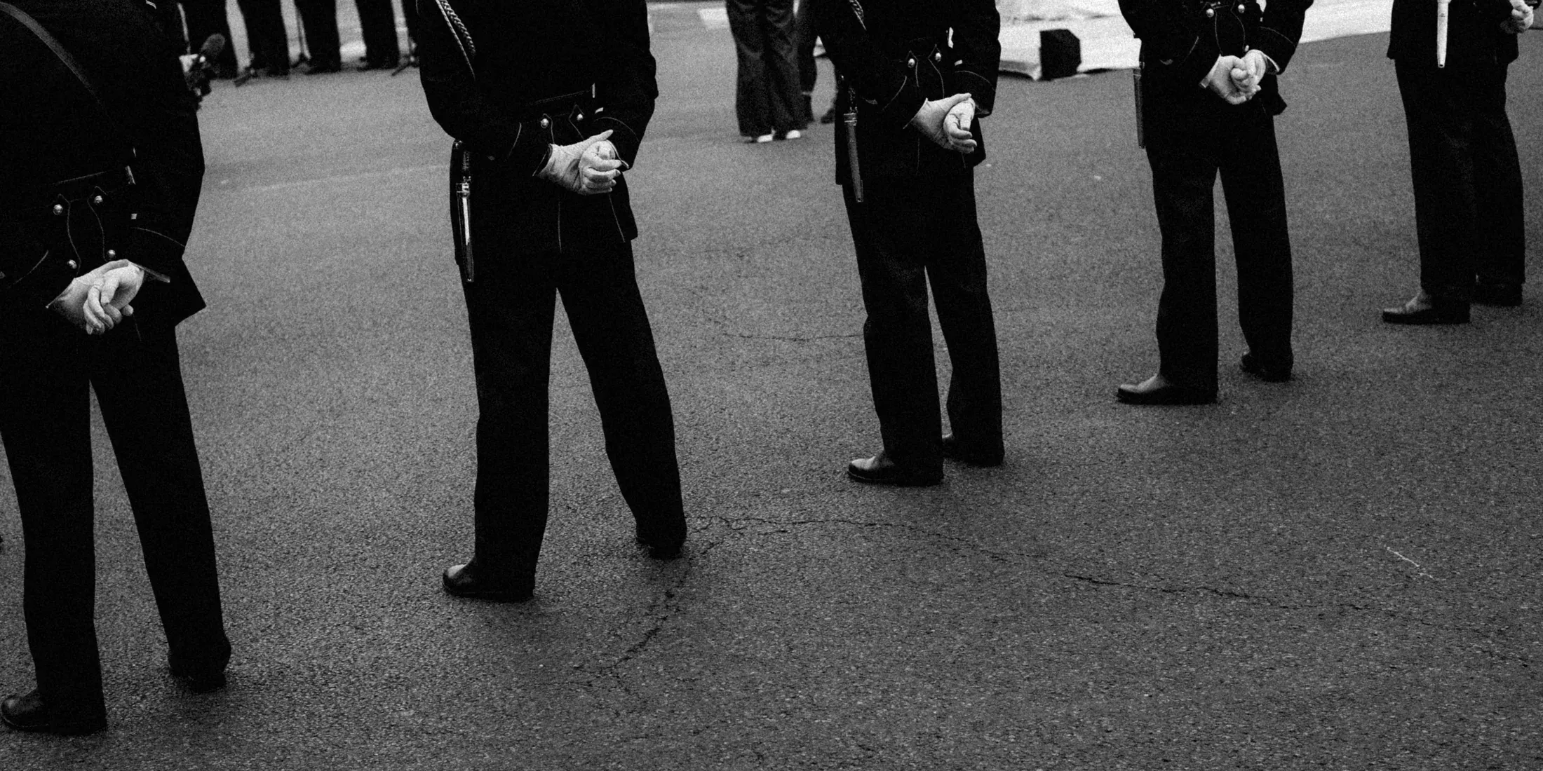 Black and white crop of uniformed figures standing in a loose circle with hands clasped.
