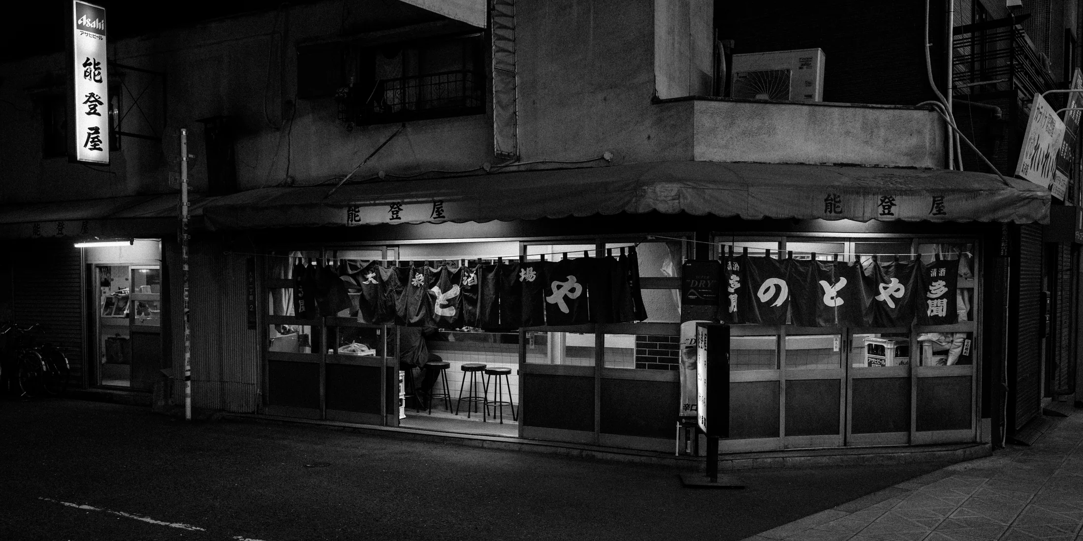 Black and white night storefront with a single figure standing at the corner.